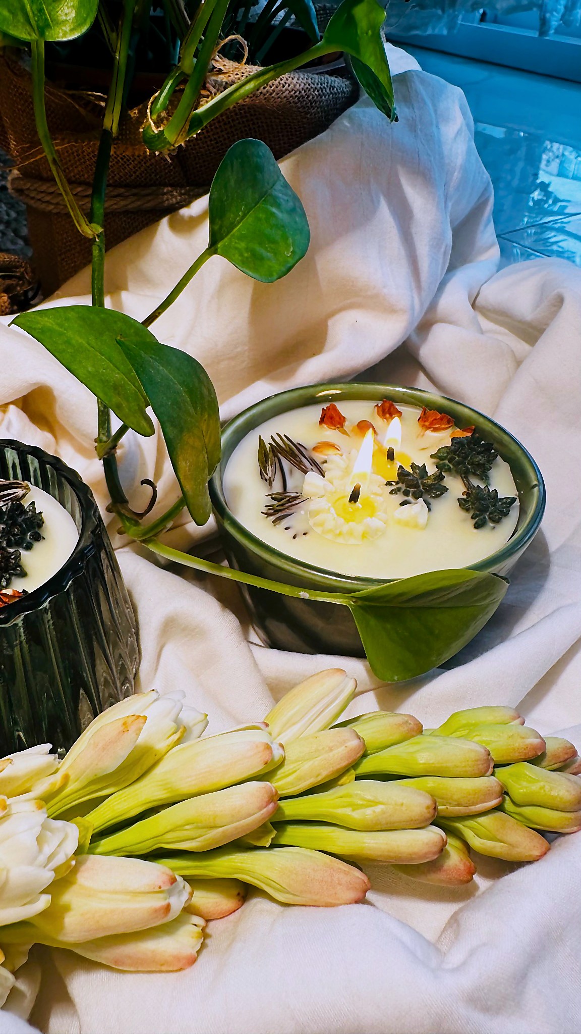A green glass vessel candle, lit, with fresh tuberose buds gathered around it